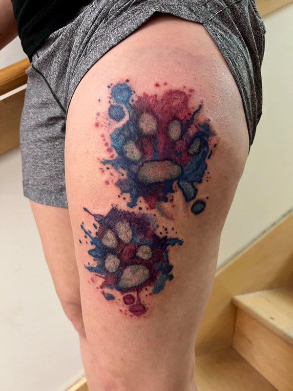 Purple, pink, and blue watercolor paw print tattoos on a woman's thigh.