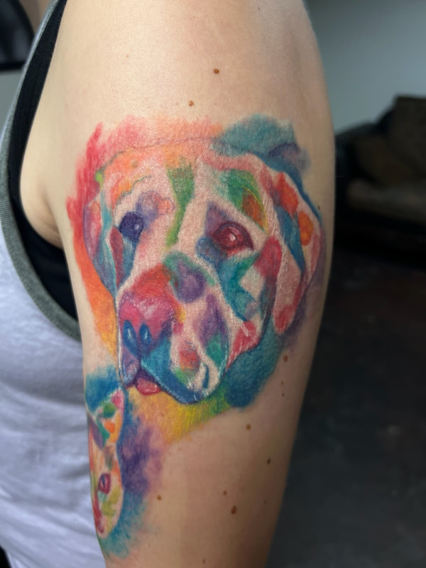 Rainbow watercolor dog face pet memorial tattoo on a woman's upper arm.