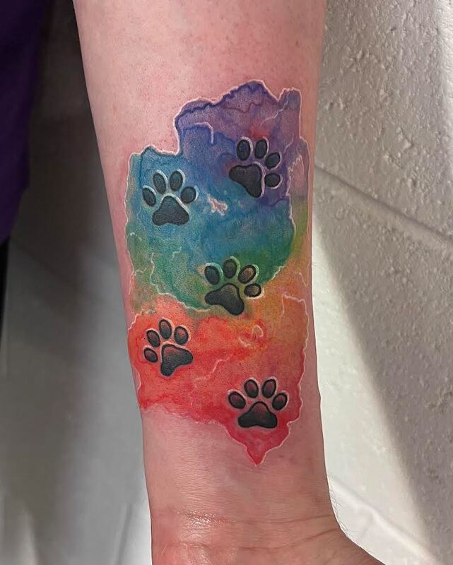 Rainbow watercolor cat paw prints tattoo on a forearm.
