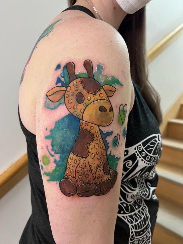 Green and blue watercolor stuffed giraffe tattoo on an outer arm.